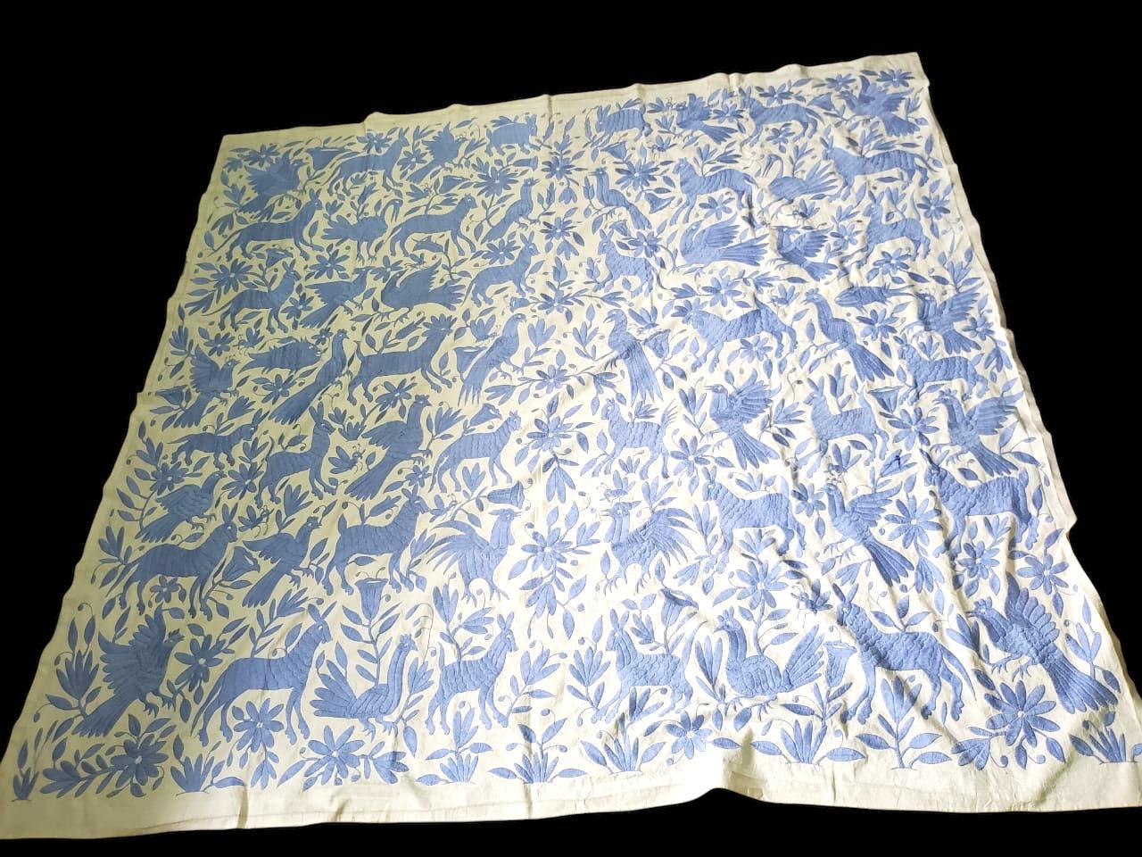 A pale blue Otomi tapestry featuring hand-embroidered patterns with a floral and animal motif .