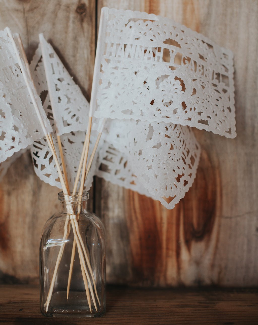 White tissue paper waving flags with cut-out patterns, attached to wooden sticks, displayed in a clear glass jar against a wooden background.