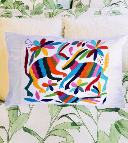 A multicolored cushion cover with an intricate traditional Otomi animal and flower motif, displayed on a cushion with a green leafy pattern in the background.