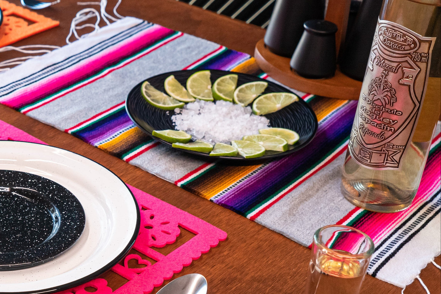 Mexican table setting with plates, lime wedges, a bottle of tequila on a colourful Mexican serape placemat.
