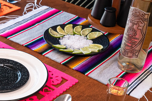 Mexican table setting with plates, lime wedges, a bottle of tequila on a colourful Mexican serape placemat.