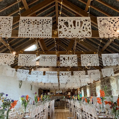 Mexican wedding papel picado banners and string lights hanging from a rustic wooden barn ceiling, with decorated tables and chairs below.