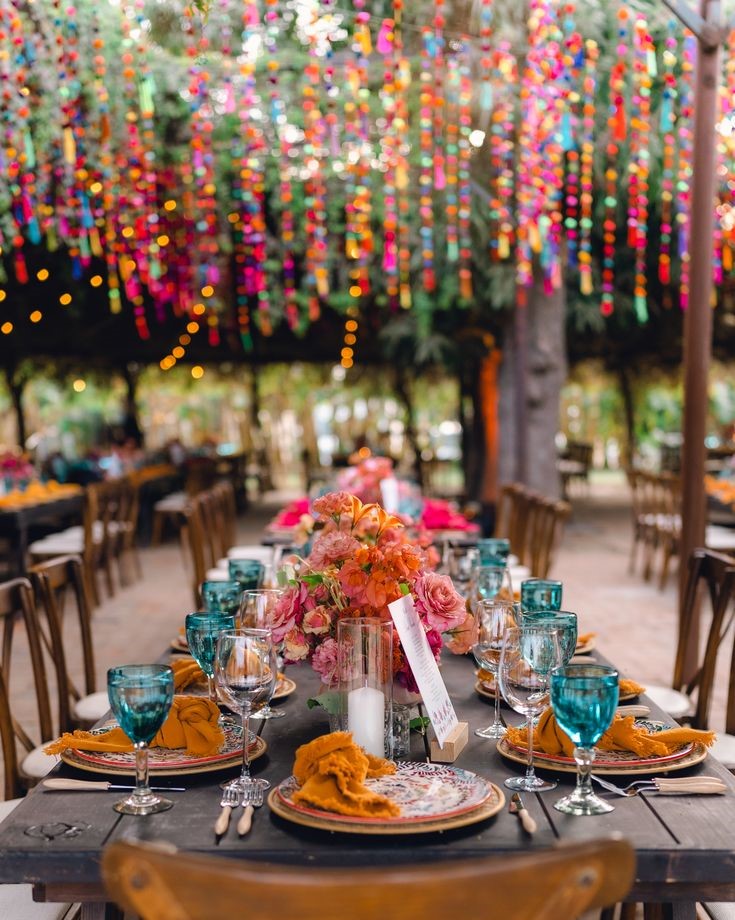 Decorative table setting with colorful flowers and glasses under a vibrant hanging pom pom garland installation.