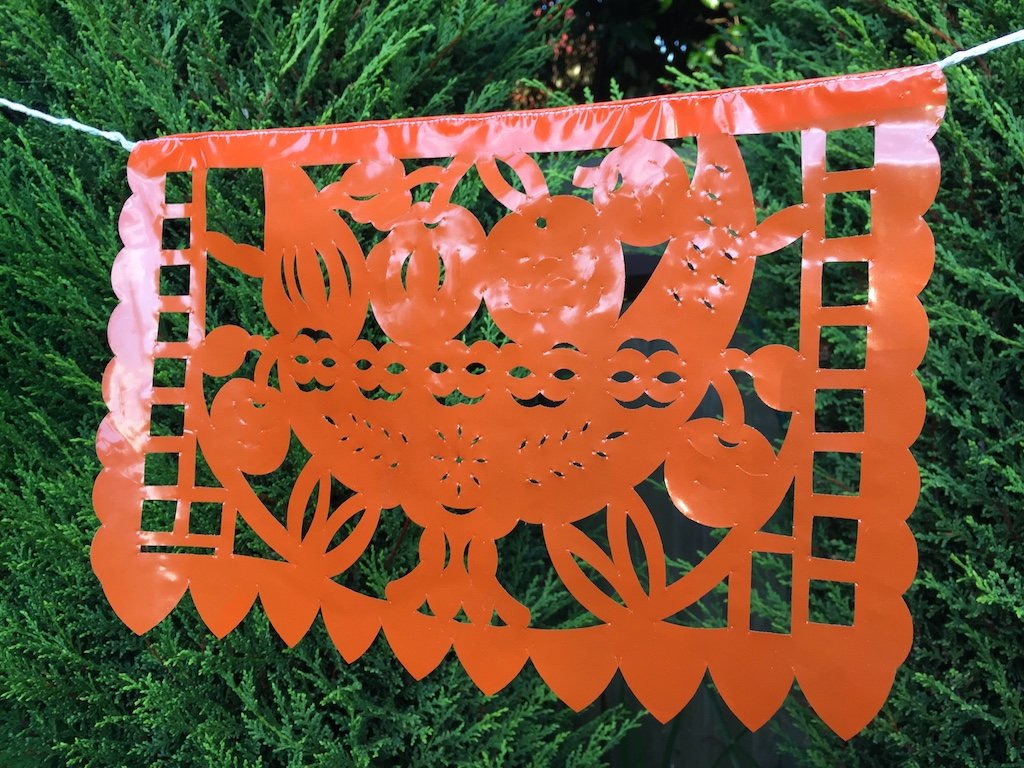 A colorful intricately cut orange plastic Mexican flag hanging on a rope, depicting a fruit bowl, decorating an outdoor garden party.