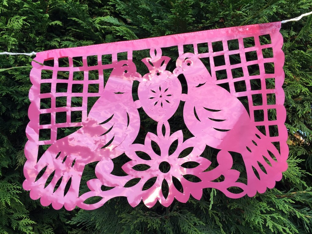A colorful intricately cut pink plastic Mexican flag hanging on a rope, depicting two birds, decorating an outdoor garden party.