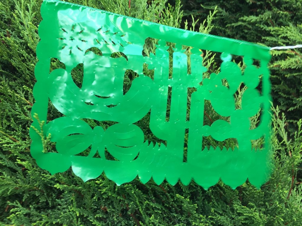 A colorful intricately cut green plastic Mexican flag hanging on a rope, depicting a cactus and a nopal, decorating an outdoor garden party.