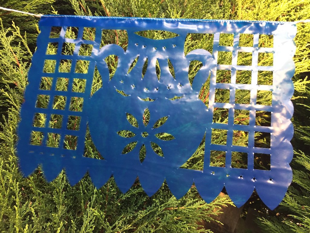 A colorful intricately cut dark blue plastic Mexican flag hanging on a rope, depicting a pot, decorating an outdoor garden party.